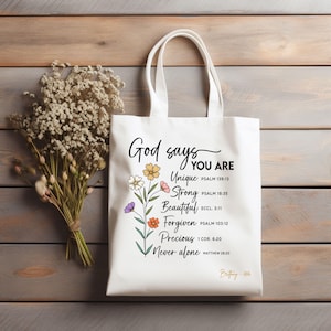 Puede incluir: Bolso tote blanco con el texto "God says YOU ARE Unique, Strong, Beautiful, Forgiven, Precious, Never alone." Diseño floral en amarillo, rosa, morado y naranja. El bolso tiene dos asas.
