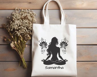 May include: White tote bag featuring a black silhouette of a person in a yoga pose, with lotus flower designs. The name "Samantha" is printed below the figure. The bag has two handles and is set against a wooden background.