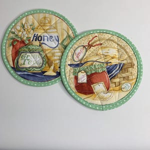May include: Two round quilted pot holders with a green polka dot border. One features a honey pot, the words "Honey" and "Green Tea". The other has a jar of jam, a basket of scones, and the word "Scones".