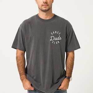 May include: Dark gray t-shirt with the text "DANCE Dads CLUB" in a white, curved design on the upper left chest. The shirt has a vintage, worn look and a crew neck.