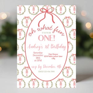 May include: A pink and white birthday invitation with the text "oh what fun IT IS TO BE ONE!" and "Audrey's 1st Birthday". The invitation has a pink oval frame and a pattern of wreaths and nutcrackers.
