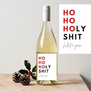 Könnte beinhalten: Eine Flasche Weißwein mit einem Etikett mit der Aufschrift "HO HO HOLY SHIT What a year!" in Rot und Schwarz. Eine Karte mit dem gleichen Text befindet sich neben der Flasche. Stechpalmen und Tannenzapfen sind im Vordergrund angeordnet.