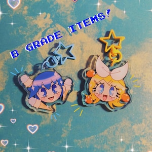 May include: Two colorful cartoon character charms with star-shaped keychains. The charms are made of clear plastic and feature a blue-haired character with a white ice cream cone and a yellow-haired character with a pink bow.