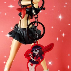 May include: A red and black anime character keychain with a black chain and a silver ring. The keychain features a female character with red hair and a red and black outfit holding a sword.