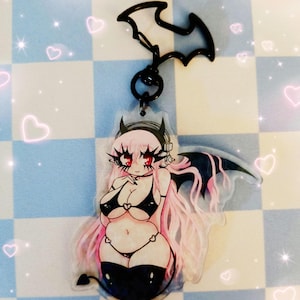 May include: A clear acrylic charm featuring a cartoon illustration of a female figure with pink hair, red eyes, and black attire. The charm has a black bat-shaped clasp and a black ring. The background is a blue and white checkered pattern with pink heart accents.