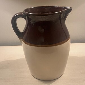 May include: A ceramic pitcher with a dark brown top and handle, a tan band, and a cream-colored base. The pitcher has a spout for pouring and a curved handle for easy grip. This vintage-style pitcher is ideal for serving beverages.