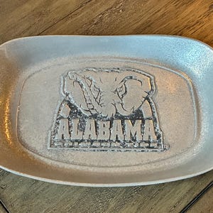 May include: A silver-colored, rectangular tray with rounded corners. The tray features a raised emblem of an elephant and the word "ALABAMA" in capital letters. The tray has a textured surface and is likely made of metal.