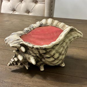 May include: A seashell-shaped decorative bowl with a pink interior. The bowl is made of ceramic with a textured, black and white exterior. The bowl has three small feet and is sitting on a wooden surface. The bowl is empty.