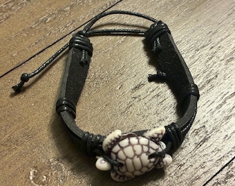 May include: Black leather bracelet featuring a white and brown turtle charm. The bracelet has a braided leather design and an adjustable cord closure. The turtle charm is detailed with a patterned shell.