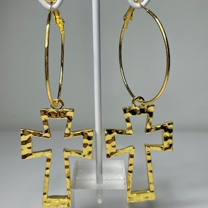 May include: Gold-coloured hoop earrings featuring a hammered cross pendant. The earrings are displayed on a white stand against a light background. The cross pendants have a textured surface and a cutout design.