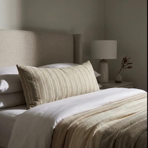 May include: A bedroom interior with a bed featuring a beige headboard, white sheets, and a striped pillow. A beige and white striped blanket is draped over the bed. A white lamp and vase with a plant are on the bedside table.