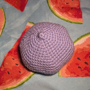 May include: A small, lavender crocheted ball with a textured surface. The ball is set against a light blue background with a watermelon pattern in red, pink, and green.