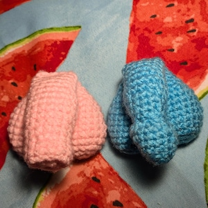 May include: Two handmade crochet objects, one pink and one blue, resembling testicles. The items are placed on a light blue fabric with a watermelon slice pattern. The crochet work is visible, showing the texture of the yarn.