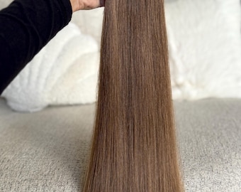 以下が含まれることがあります： 茶色のヘアエクステンションの束を手に持っています。エクステンションはミディアムブラウンで、ストレートに見えます。髪は滑らかで光沢があります。背景はぼやけており、白い枕とテクスチャのある表面があります。