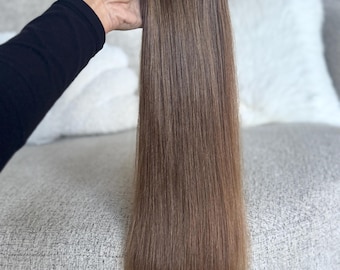 以下が含まれることがあります： 明るい茶色のヘアエクステンションの束。エクステンションは黒い袖の手に持たれています。髪はストレートで、長さは約50cmです。背景は明るい色のソファと枕です。