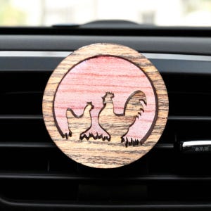 May include: A round wooden car air freshener featuring a rooster and hen design. The air freshener has a light brown outer ring and a pinkish-red center. The rooster and hen are cut out of the wood, creating a silhouette.