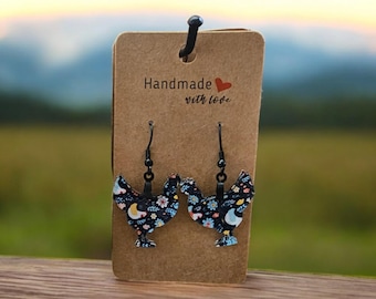 May include: A pair of black chicken-shaped earrings with a floral pattern. The earrings are hanging on a brown card with the text "Handmade with love".
