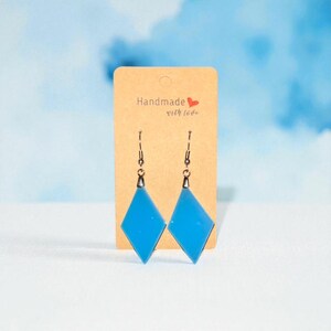 Peut inclure: Une paire de boucles d'oreilles faites à la main en forme de losange bleu avec des crochets noirs, présentées sur une carte beige avec le texte "Handmade with love". Le fond est bleu clair avec des accents blancs.