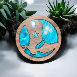 May include: Round wooden coaster with a blue resin design. The coaster features a crescent moon shape with a star and a sun-like element. The wood has a natural grain, and the resin has a swirling, ocean-like appearance.