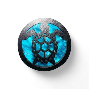 May include: A circular bottle stopper with a black, textured exterior. It features a laser-cut geometric pattern over a vibrant, swirling blue resin. The design is reminiscent of a turtle shell.