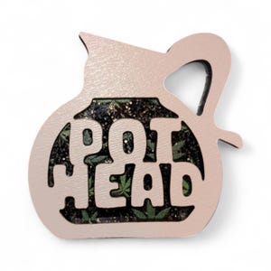 Puede incluir: Un objeto decorativo rosa claro con forma de tetera con las palabras "POT HEAD" recortadas. El interior presenta un patrón de hojas verdes con purpurina.