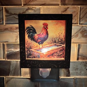 May include: A night light with a framed image of a rooster standing by a water trough. The rooster is depicted with red, blue, and black feathers. The night light has a black frame and is plugged into a wall socket.