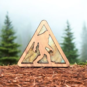 Puede incluir: Letrero de Bigfoot de madera en forma triangular, con una silueta recortada de la criatura. El letrero tiene un marco de madera marrón claro y un centro relleno de resina con tonos dorados y beige brillantes. El fondo muestra un bosque.