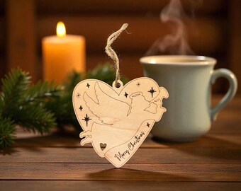 May include: Wooden heart-shaped Christmas ornament with an angel design and the words "Merry Christmas". The ornament is suspended by a twine string. A lit candle and a mug of coffee are in the background.