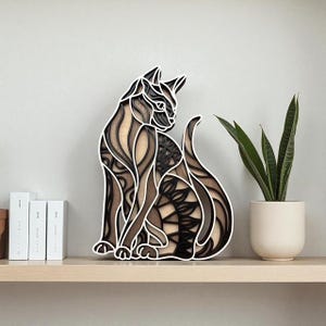 May include: A layered wooden cat sculpture in shades of brown, black, and white. The cat is in a seated position with its tail curled. The artwork is placed on a light-colored shelf with a potted plant and books in the background.