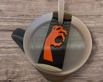 May include: A Halloween-themed drink stirrer featuring an orange hand design on a black background. The stirrer is inserted into a clear cup with a dark handle. The cup is filled with a clear liquid, and the stirrer is made of plastic.