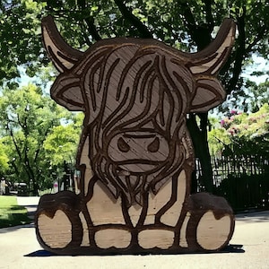 Könnte beinhalten: Eine Holzskulptur einer schottischen Hochlandkuh, braun und weiß bemalt, mit detailliertem Gesicht und Hörnern. Die Skulptur sitzt auf einem Weg in einem Park.