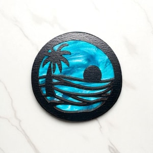 May include: A circular black coaster with a tropical sunset design. It features a palm tree silhouette, a sun, and layered lines depicting the sea. The background is a vibrant mix of blue and white, evoking the sky and water.