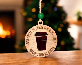 Puede incluir: Adorno navideño redondo de madera con el texto "Dear Santa I'm sorry for what I said before I had my coffee". Presenta un diseño de taza de café y está suspendido de una cuerda con una cuenta verde.