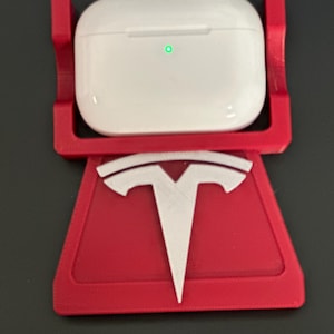 Puede incluir: Un estuche de carga de auriculares inalámbricos blanco descansa sobre un soporte rojo con un logotipo blanco de Tesla. El soporte está diseñado para sujetar el estuche de carga. El estuche de carga tiene una luz verde.