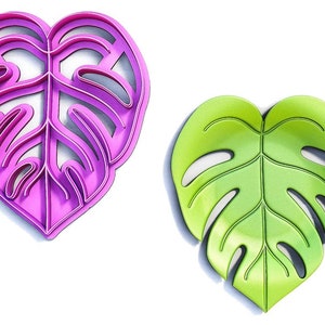 Puede incluir: Se muestran dos objetos con forma de hoja, uno rosa brillante y otro verde, sobre un fondo blanco. La hoja rosa es un cortador de galletas, mientras que la hoja verde parece ser un producto terminado. Ambos tienen un diseño de hoja de monstera.