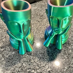 Op de afbeelding: Twee regenboogkleurige groene keramische plantenpotten in de vorm van mensenhoofden met hun kin op hun handen rustend. De potten hebben een gladde, glanzende afwerking en zijn perfect voor het tentoonstellen van kleine planten of bloemen.