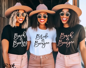 Op de afbeelding: Drie vriendinnen dragen zwarte, witte en zwarte T-shirts met de tekst "Bach & Boozy", "Bride & Boujee" en "Bach & Boujee" in zwarte en roségouden letters. De vriendinnen dragen hoeden en zonnebrillen.