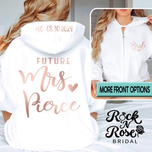 May include: White zippered hoodie with the text "Future Mrs. Pierce" in rose gold lettering on the back and "Bride" in rose gold lettering on the front. The hoodie is worn by a person with long blonde hair. The text "Rock N Rose Bridal" is visible at the bottom of the image.