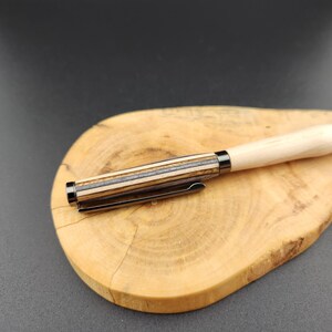 May include: A wooden pen with a light-colored barrel and a dark, striped section near the tip. The pen has a metal clip and is resting on a wooden cutting board with a natural wood grain pattern.