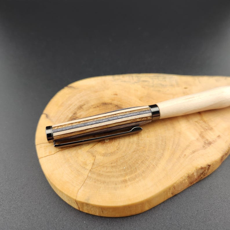 May include: A wooden pen with a light-colored barrel and a dark, striped section near the tip. The pen has a metal clip and is resting on a wooden cutting board with a natural wood grain pattern.