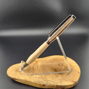 May include: A handcrafted pen with a light wood barrel and a multi-toned wood and metal cap. The pen is displayed on a wooden base and clear acrylic stand, showcasing its design and craftsmanship. The pen's tip and clip are silver.