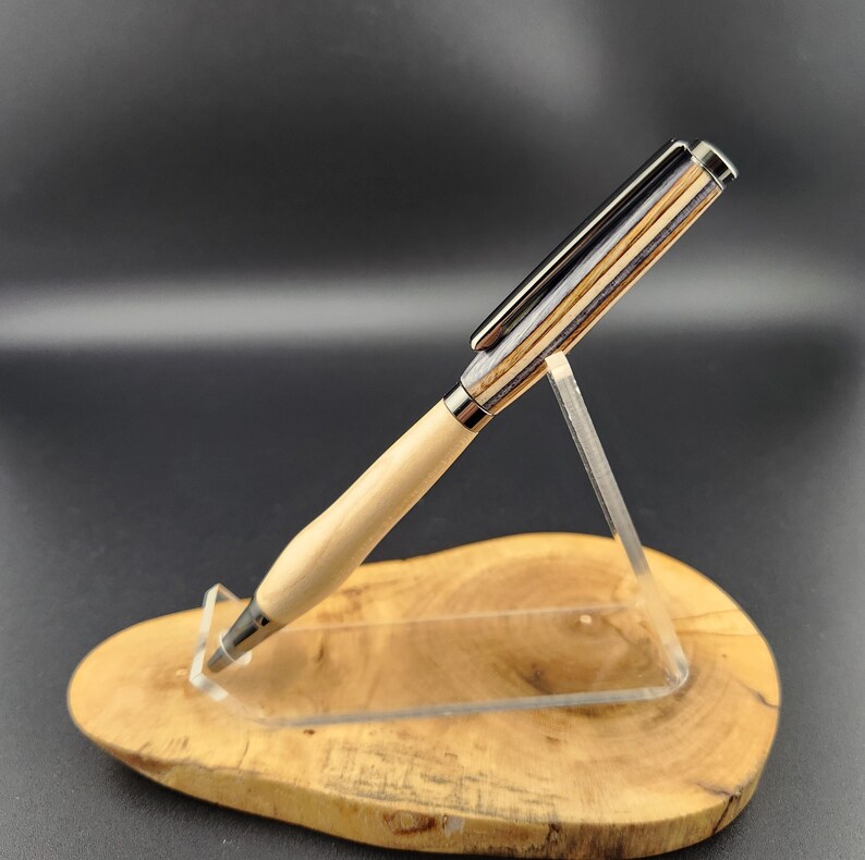 May include: A handcrafted pen with a light wood barrel and a multi-toned wood and metal cap. The pen is displayed on a wooden base and clear acrylic stand, showcasing its design and craftsmanship. The pen's tip and clip are silver.