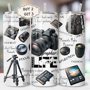 May include: A white tumbler with a black and white design featuring a camera, a tripod, and text related to photography. The text includes "Photographer's Life", "Moments Make Memories", "Every Picture Tells a Story", and more. The tumbler has a black lid and a straw.