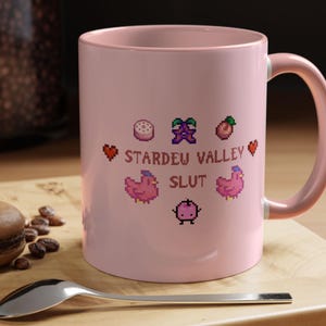 Peut inclure: Une tasse en céramique rose clair avec une anse assortie. La tasse présente des graphiques pixélisés et le texte "Stardew Valley Slut". Le design comprend des cœurs, un poulet et d'autres icônes liées au jeu. Une cuillère et un macaron sont au premier plan.