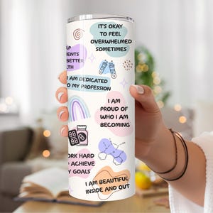 May include: A white tumbler with a pink, blue, and purple design. It features motivational quotes like "It's okay to feel overwhelmed sometimes" and "I am dedicated to my profession".