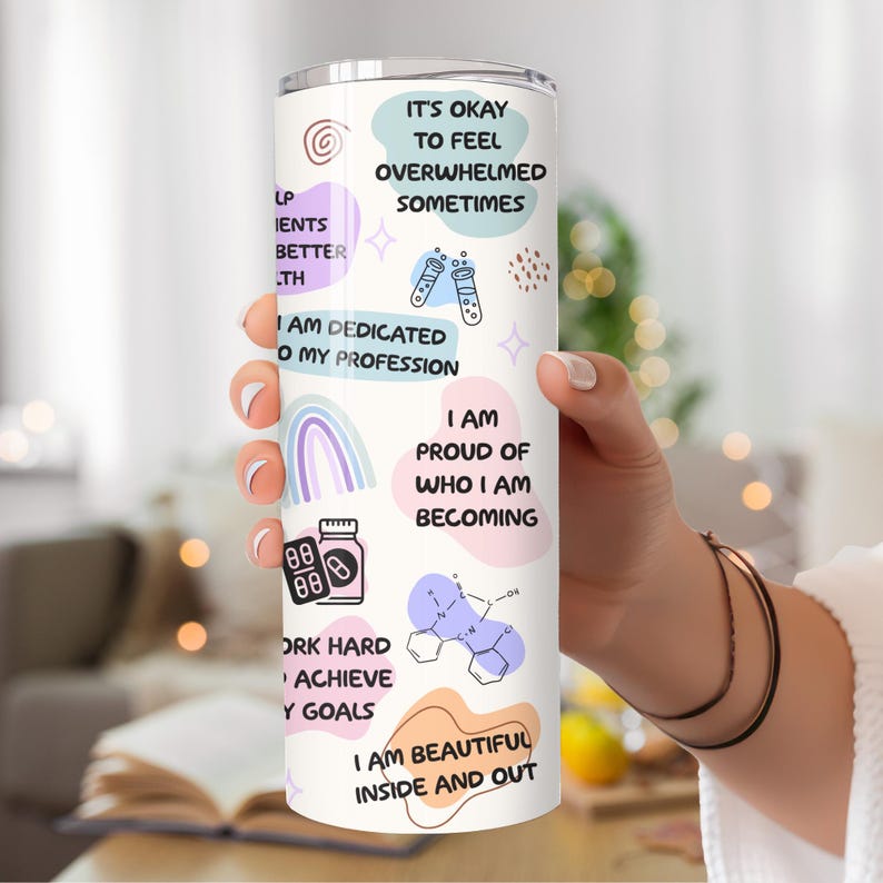 May include: A white tumbler with a pink, blue, and purple design. It features motivational quotes like "It's okay to feel overwhelmed sometimes" and "I am dedicated to my profession".