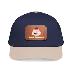 May include: A navy blue baseball cap with a beige peak. The cap has a brown leather patch with a cartoon fox character wearing a tomato. The text "Team Tomafox" is printed on the patch.