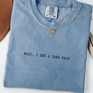 May include: A blue t-shirt with the text "WAIT, I SEE A YARD SALE" in black. A gold necklace with a pendant is draped over the shirt. A brown hat and dried plant are in the background.