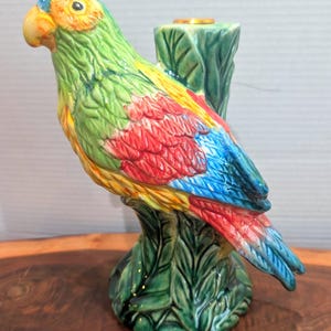 May include: A colorful ceramic parrot figurine perched on a green tree trunk base. The parrot features vibrant green, yellow, red, and blue plumage. The figurine has a gold-colored ring on top of the tree trunk.