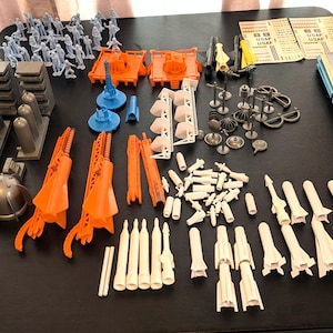 May include: A collection of vintage toy military and space-themed components. Includes miniature soldiers, orange launch platforms, missiles, and various other plastic pieces in shades of gray, orange, blue, and white. A nostalgic set for collectors.